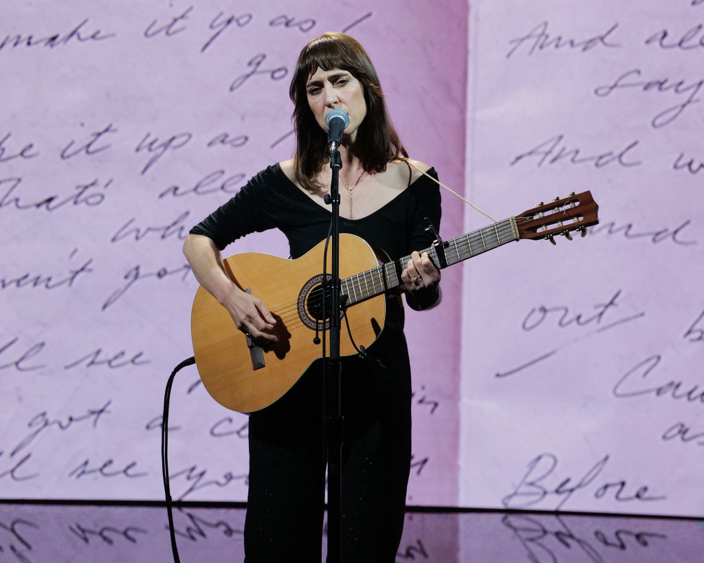 Leslie Feist is wearing black and playing an acoustic guitar against a pink background