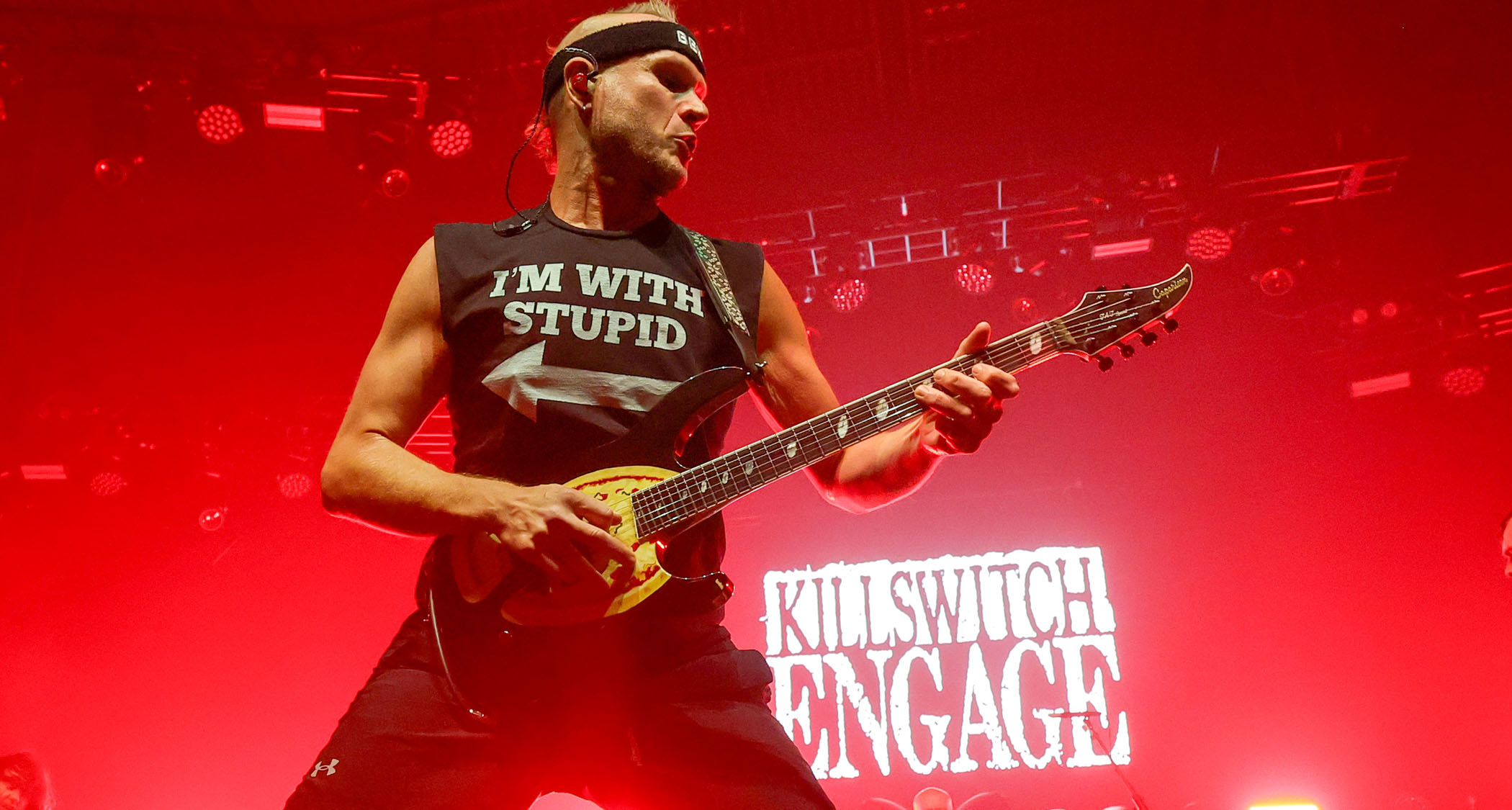 Adam Dutkiewicz onstage with Killswitch Engage, wearing a black sweatband and sleevless T-shirt, red lights and the band's logo are in the background.