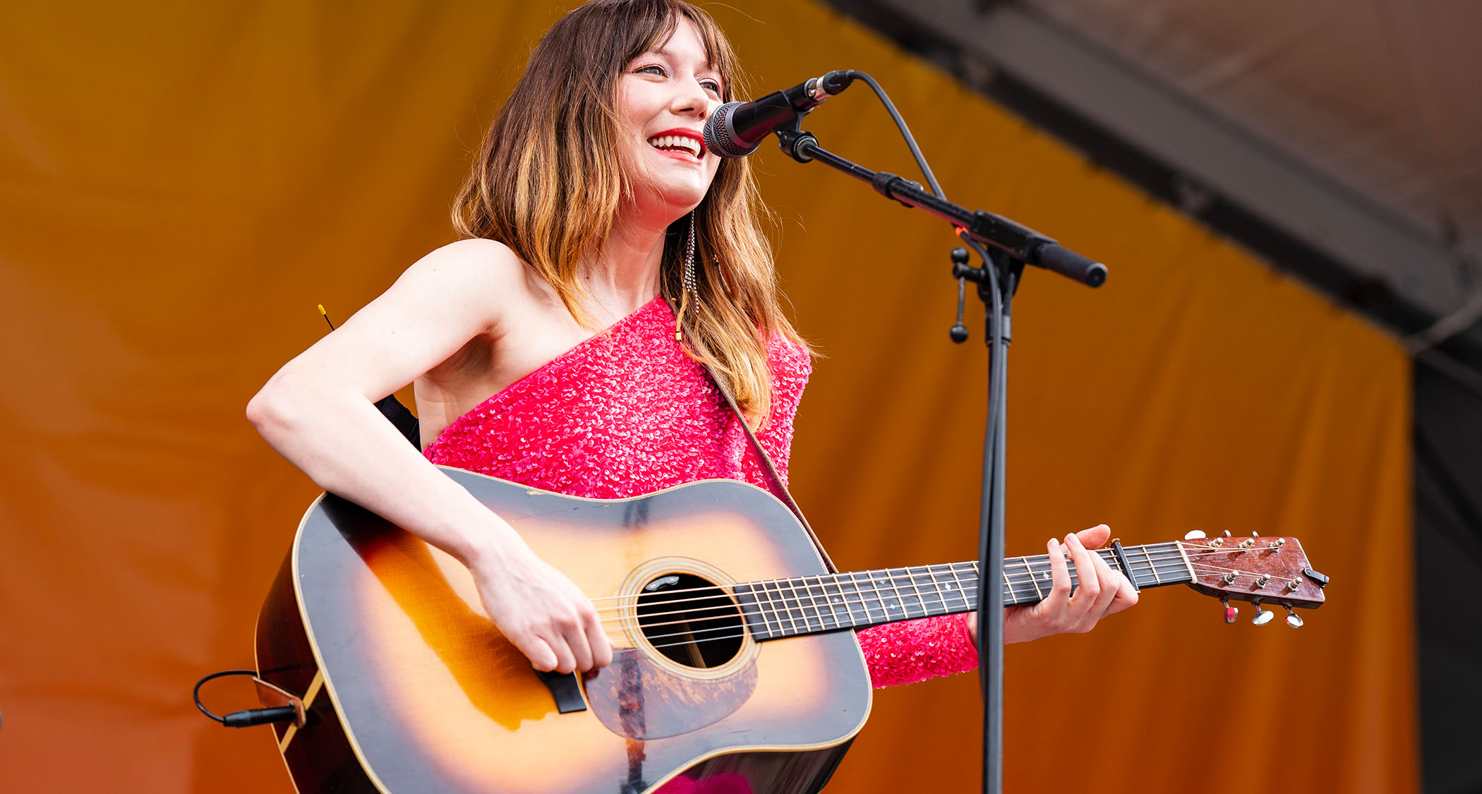 Molly Tuttle plays a sunburst dreadnought onstage and wears a pink sparkly dress as she performs at the 2024 New Orleans Jazz &amp; Heritage Festival.